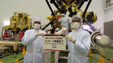 Deux personnes en tenue blanche, portant gants, charlottes et masques tiennent un panneau avec des inscriptions en japonais et la mention "message from the Earth installation complete - JAXA".