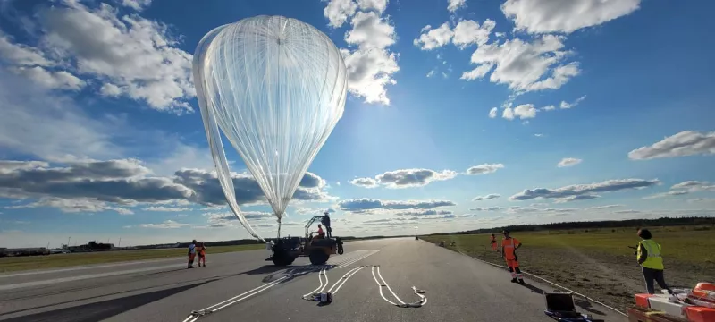 Un homme conduit un véhicule retenant un ballon au sol. Au loin, plusieurs personnes autour du véhicule.